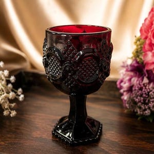 Könnte beinhalten: Ein weinrotes Weinglas im Vintage-Stil mit aufwendigen Details. Das Glas hat ein strukturiertes Muster und einen quadratischen Fuß. Das Glas steht auf einer dunklen Holzoberfläche, mit Blumen im Hintergrund.