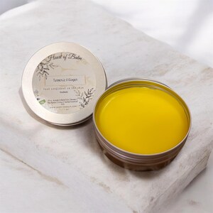 May include: A round, silver tin of turmeric and ginger balm with the lid off, revealing a bright yellow balm. The lid is next to the open tin, with the text "Heart of Balm" and "Turmeric & Ginger" printed on it. The balm is handmade.