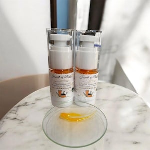 May include: Two white and clear cosmetic bottles with orange liquid and white pumps, labeled "Heart of Balm". A small glass dish contains a smear of the same orange liquid. The bottles are on a marble surface.