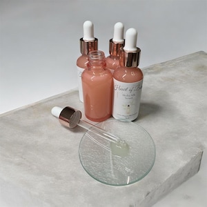 May include: Four pink serum bottles with white labels and rose gold droppers are arranged on a gray surface. One bottle is open with a dropper and a small glass dish. The labels read "Heart of Bliss Hydra Silk Serum".