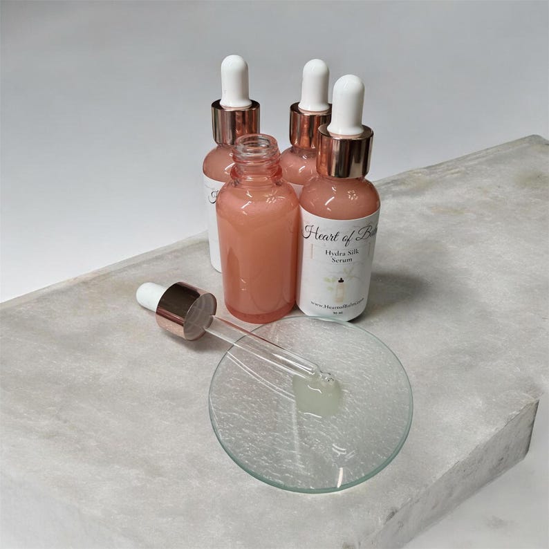 May include: Four pink serum bottles with white labels and rose gold droppers are arranged on a gray surface. One bottle is open with a dropper and a small glass dish. The labels read "Heart of Bliss Hydra Silk Serum".
