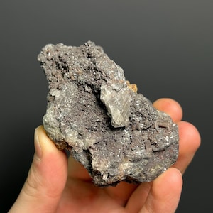 Könnte beinhalten: Nahaufnahme einer Mineralprobe, die in einer Hand gehalten wird. Der Stein hat eine raue, strukturierte Oberfläche mit einer Mischung aus dunkelgrauen, braunen und hellgrauen Farben. Einige kristalline Strukturen sind sichtbar.