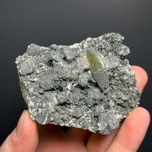 May include: A rough, gray rock formation with a prominent, elongated, olive-green crystal protruding from its surface. The crystal has a pointed tip and a translucent appearance. The rock has a textured, granular surface. The background is a dark gray.