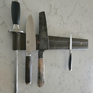 May include: A collection of kitchen knives and a sharpening steel are mounted on a dark green magnetic knife holder. The knives have various blade shapes and handle designs, including wood and black handles.