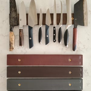 May include: A set of ten knives, including a cleaver, are displayed on a magnetic knife rack. The rack is made of brown leather and has four additional shelves in brown, gray, and black.