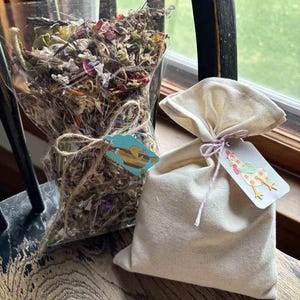 May include: A bouquet of dried flowers in a clear plastic wrap tied with twine, and a small beige burlap bag with a tag. The tag has a drawing of a chicken. The items are on a wooden surface.