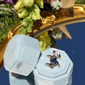 May include: Two gold-toned rings featuring wizard hat designs with enamel accents, one in light pink and one in dark blue, each decorated with small star and crescent moon symbols, displayed inside a light blue velvet octagonal ring box.