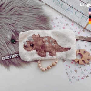 Puede incluir: Una bolsa blanca de crochet con un diseño de triceratops tejido en marrón. La bolsa está sobre un fondo blanco con estrellas rosas y azules.