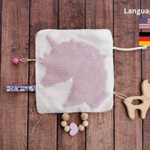 Puede incluir: Un juguete de bebé de punto rosa y blanco con un diseño de unicornio. El juguete tiene una llama de madera unida a él con una cuerda. El juguete también tiene un corazón de madera rosa y una cadena de cuentas de madera.