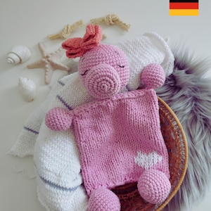 Könnte beinhalten: Ein rosa gehäkeltes Stofftier mit einer rosa Schleife auf dem Kopf. Das Tier liegt auf einer weiß-grauen gestreiften Decke in einem Weidenkorb. Das Tier hat ein weißes Herz auf der Brust.