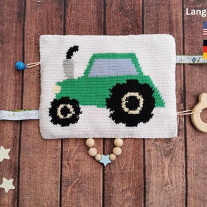 Puede incluir: Un juguete de crochet blanco para bebé con un diseño de tractor verde, un mordedor de madera y un collar de cuentas de madera con un dije de estrella azul. El juguete tiene una campana y una etiqueta con las palabras "Idiomas:" y banderas de Estados Unidos y Alemania.