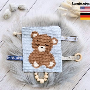 Puede incluir: Un juguete de bebé de crochet azul con un diseño de oso marrón. El juguete tiene un anillo de madera, un collar de cuentas y varias etiquetas de tela.