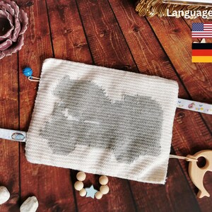 Puede incluir: Un juguete de bebé de crochet blanco con una forma de animal gris en el centro. El juguete tiene una campanilla azul, un mordedor de madera con forma de delfín y una pulsera de cuentas de madera con un dije de estrella azul.