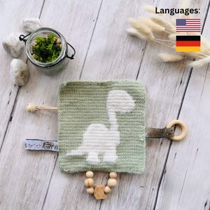 Puede incluir: Un juguete de bebé tejido a ganchillo en verde salvia con un diseño de dinosaurio blanco. El juguete tiene un anillo de madera, una cuenta de madera en forma de estrella y una pequeña campana. Se adjunta una pequeña etiqueta de tela con ilustraciones de animales.