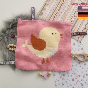 Puede incluir: Manta de bebé de crochet rosa con un diseño de pájaro amarillo y un mordedor de madera. La manta tiene una variedad de texturas y colores, incluyendo marrón, blanco y rosa.