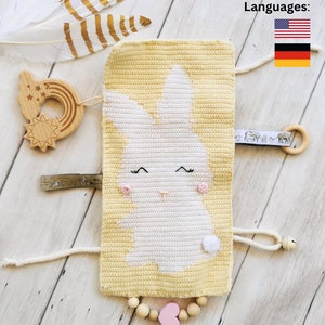 Puede incluir: Una manta de actividad para bebé de crochet amarillo con un diseño de conejo blanco. La manta tiene una variedad de texturas y características, incluyendo un sol de madera, un arcoíris, un anillo de madera, un collar de cuentas de madera y un corazón rosa.