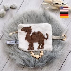 May include: A white crocheted square with a brown camel silhouette. The square is attached to a wooden ring with a brown leather strap. The square is on a gray faux fur surface. The image also includes the text "Languages:" and flags for the United States and Germany.