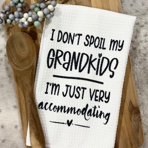 May include: White kitchen towel with the text "I DON'T SPOIL MY GRANDKIDS I'M JUST VERY accommodating" in black. The towel is on a wooden cutting board with a wooden spoon and decorative berries.