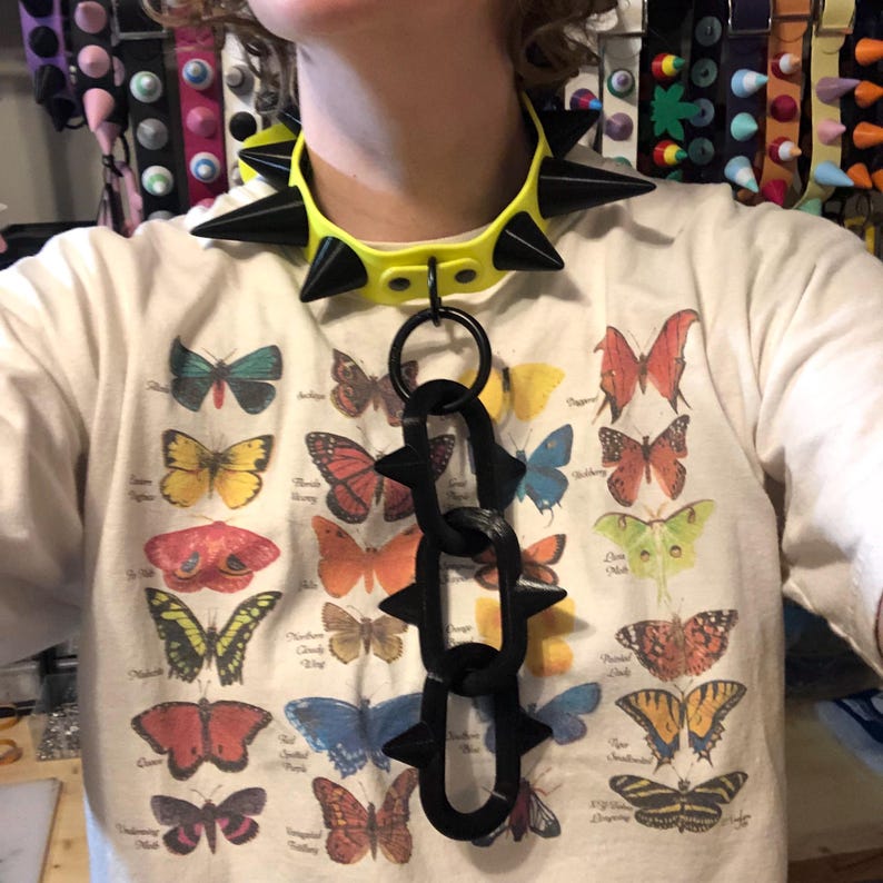 May include: A bright yellow choker collar featuring large black spikes and a dangling chain attachment with three black spiked links. The collar is worn over a cream-colored t-shirt printed with a grid of various colorful butterfly illustrations.