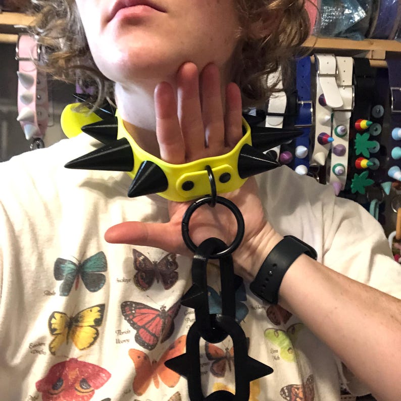 May include: A bright neon yellow collar featuring large, pointed black spikes and a central black O-ring. Attached to the O-ring is a vertical chain composed of two black oval links, with the lower link also featuring two black spikes on its sides.