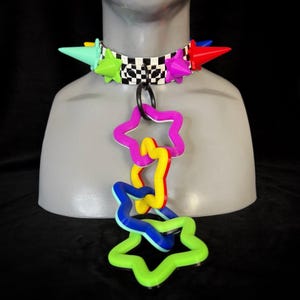 May include: A black and white checkered choker collar featuring multi-colored cone-shaped spikes in mint, lime, purple, and red. A black metal ring at the center holds a dangling chain of five interlocking star-shaped links in purple, yellow, blue, and lime.