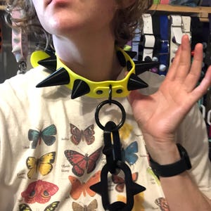 May include: A bright neon yellow collar featuring large, pointed black spikes and a central black O-ring. Attached to the O-ring is a vertical chain of three black, elongated oval links, with the bottom link also featuring two black spikes.