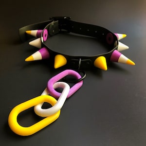 May include: Black leather dog collar with colorful, cone-shaped spikes in yellow, purple, and white. The collar has a buckle closure and a metal ring with a chain of yellow, purple, and white links.
