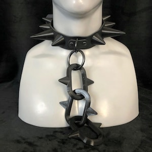 May include: Black leather collar with large, pointed spikes. A matching chain with spike accents hangs from the collar. The collar is displayed on a white mannequin bust, showcasing its design and construction.