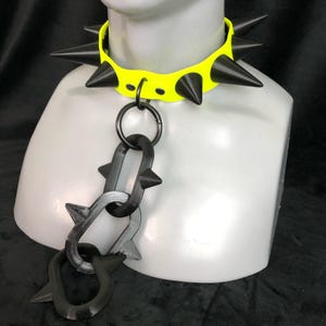 May include: A neon yellow choker collar featuring large black spikes along the band. A central metal ring connects to a dangling chain composed of three interlocking black links, each adorned with smaller spikes, displayed on a mannequin neck.