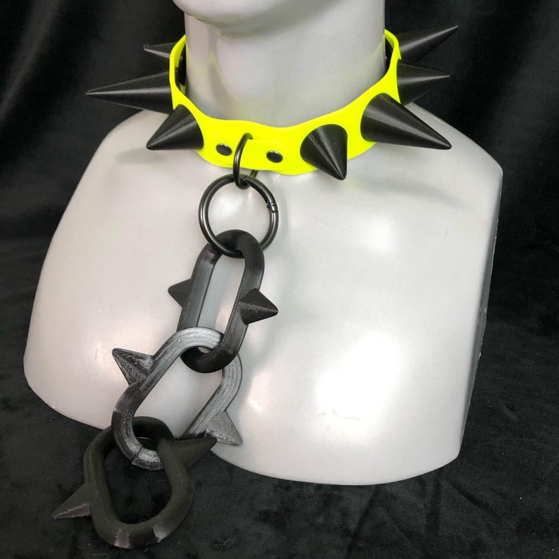 May include: A neon yellow choker collar featuring large black spikes along the band. A central metal ring connects to a dangling chain composed of three interlocking black links, each adorned with smaller spikes, displayed on a mannequin neck.