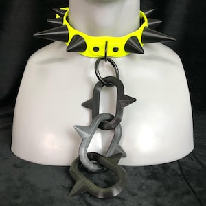 May include: A neon yellow collar featuring multiple sharp black spikes along the band. A large metal ring is attached to the front, connecting to a vertical chain composed of three interlocking black links, each featuring small protruding spikes.