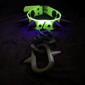 May include: A neon yellow collar featuring black conical spikes and a matching black chain attachment with spiked links. The collar includes a buckle closure and circular black rivets securing the spikes to the vibrant yellow band material.