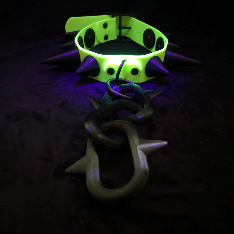 May include: A neon yellow collar featuring black conical spikes and a matching black chain attachment with spiked links. The collar includes a buckle closure and circular black rivets securing the spikes to the vibrant yellow band material.