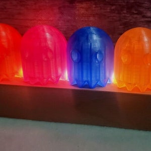 Pode incluir: Quatro lâmpadas em forma de fantasma com abajures de plástico translúcido nas cores vermelho, rosa, azul e laranja. As lâmpadas estão dispostas em uma fileira sobre uma base de madeira.