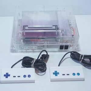 Może przedstawiać: Przezroczysta konsola Nintendo Entertainment System (NES) z dwoma białymi kontrolerami. Kontrolery mają niebieskie pady kierunkowe i przyciski. Konsola ma przezroczystą plastikową obudowę.