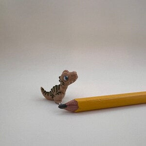 Könnte beinhalten: Eine kleine, braune, 3D-gedruckte Dinosaurierfigur mit schwarzen Streifen und blauen Augen. Der Dinosaurier steht auf einer weißen Oberfläche neben einem gelben Bleistift.