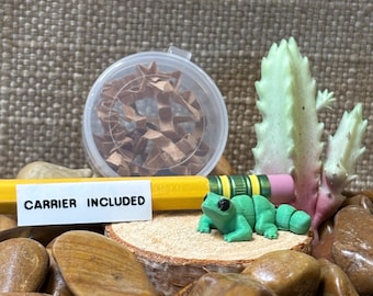 May include: A small green articulated frog figurine sits on a wooden slice next to a yellow pencil with a pink eraser. A clear plastic container filled with brown paper shreds is behind the pencil. A white label reads CARRIER INCLUDED.