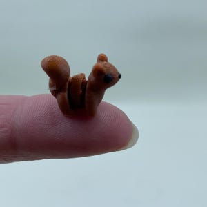 Könnte beinhalten: Eine Miniatur-Figur eines braunen Eichhörnchens mit einem eingerollten Schwanz und schwarzen Augen. Die kleine Skulptur ruht auf einer Fingerspitze und zeigt ihre geringe Größe. Die Figur ist eine detaillierte Darstellung eines Eichhörnchens.