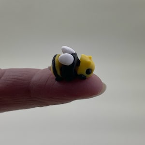 May include: A small, yellow and black bee figurine with white wings. The bee is lying on its side with its head facing the right.