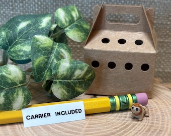 May include: A miniature brown cardboard pet carrier with ventilation holes, a yellow pencil with a pink eraser, and a tiny lion figurine. A white label on the pencil reads CARRIER INCLUDED. A green and white variegated artificial plant is visible.