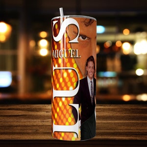 Puede incluir: Un vaso blanco con un diseño dorado y naranja que presenta un retrato de un hombre y la palabra "MIGUEL".