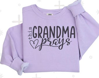 May include: A light purple sweatshirt with the text "this GRANDMA prays" in black script. The word "orchid" is written in script at the bottom. The sweatshirt is folded, showing the sleeves and the inside lining.