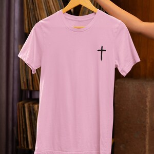 Może przedstawiać: Jasny r&oacute;żowy t-shirt z czarnym nadrukiem krzyża z przodu. Koszulka jest zawieszona na drewnianym wieszaku.
