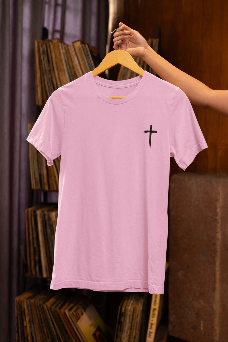 Może przedstawiać: Jasny r&oacute;żowy t-shirt z czarnym nadrukiem krzyża z przodu. Koszulka jest zawieszona na drewnianym wieszaku.