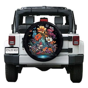 May include: A white Jeep with a spare tire cover featuring a colorful floral design on a black background. The design includes a variety of flowers, butterflies, and leaves. The text "STYLE MY WHIP TIRE COVERS & MORE" is printed on a sticker on the back of the Jeep.