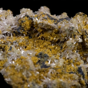 Könnte beinhalten: Nahaufnahme einer Gesteinsformation mit einem Cluster aus klaren, nadelförmigen Kristallen. Die Kristalle sind in eine Matrix aus gelben und schwarzen Mineralien eingebettet.