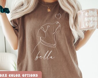 May include: A brown t-shirt with a white line drawing of a dog with the name "bella" underneath. The dog is looking up at a small heart.