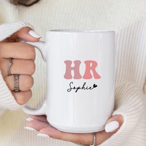 May include: White ceramic mug with a pink and red "HR" graphic and the name "Sophie" in black script. The mug has a black heart symbol next to the name.