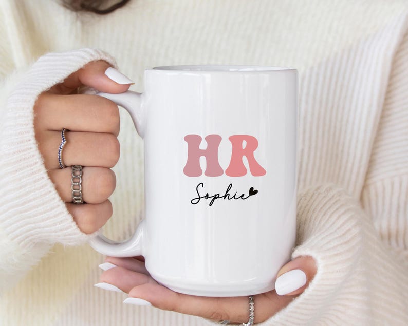 May include: White ceramic mug with a pink and red "HR" graphic and the name "Sophie" in black script. The mug has a black heart symbol next to the name.