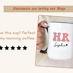 May include: White ceramic mug with a black rim and handle. The mug has pink letters that spell "HR" and the name "Sophie" written in black cursive. The mug is being held by a hand.